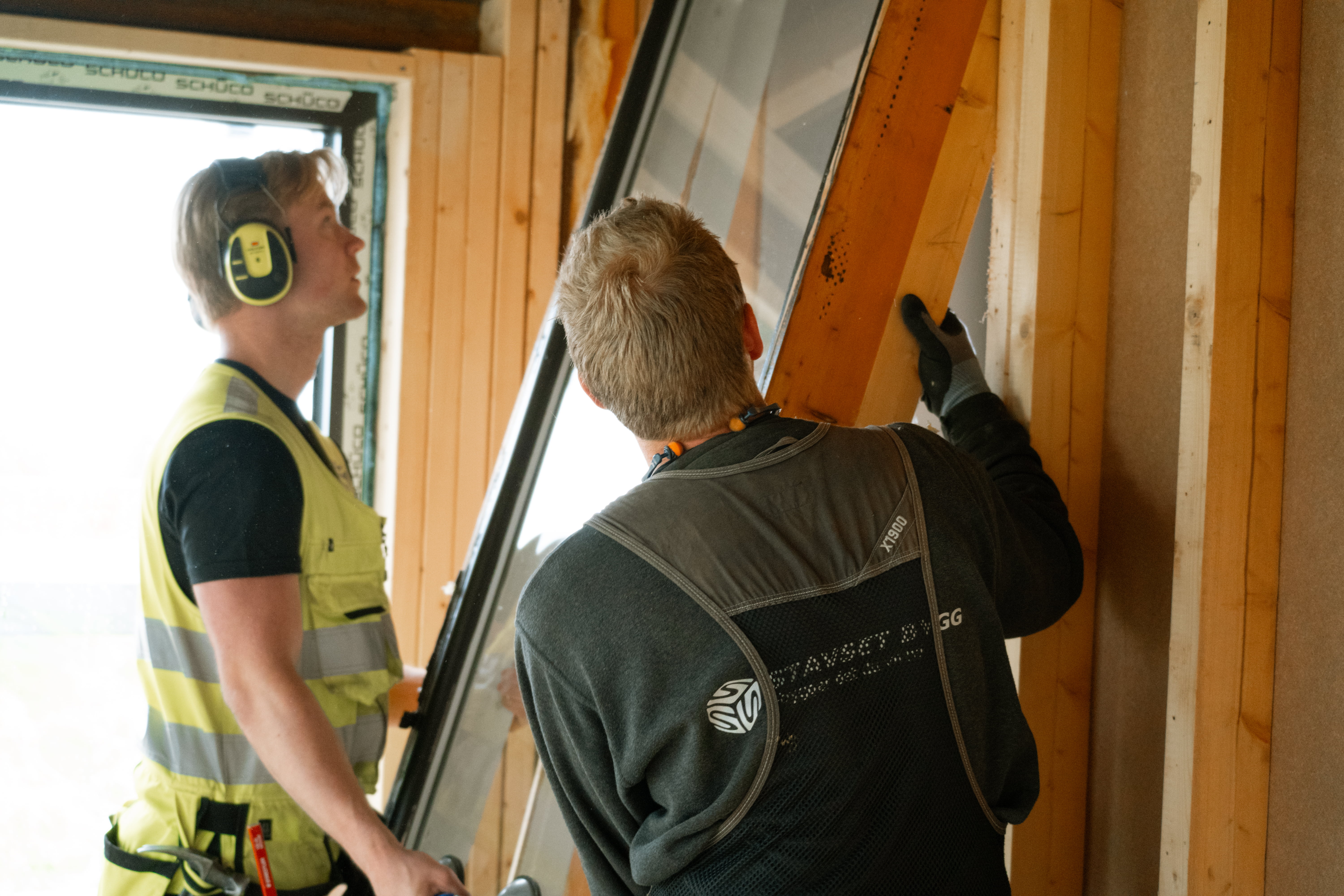 To personer monterer et stort vindu i en trekonstruksjon, med fokus på samarbeid og presisjon i arbeidet.