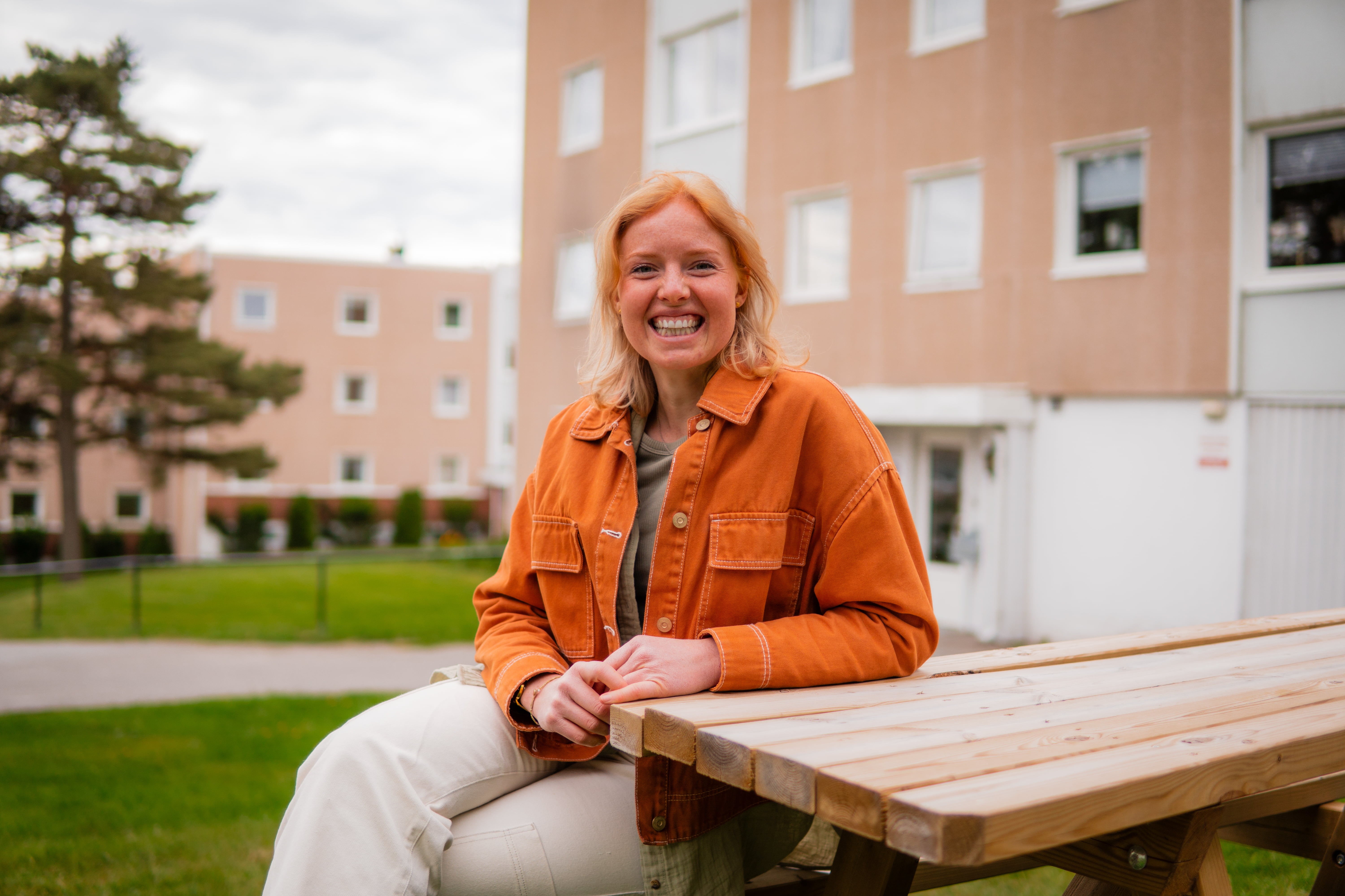 En ung, smilende kvinne sitter på en benk foran en bolig blokk.